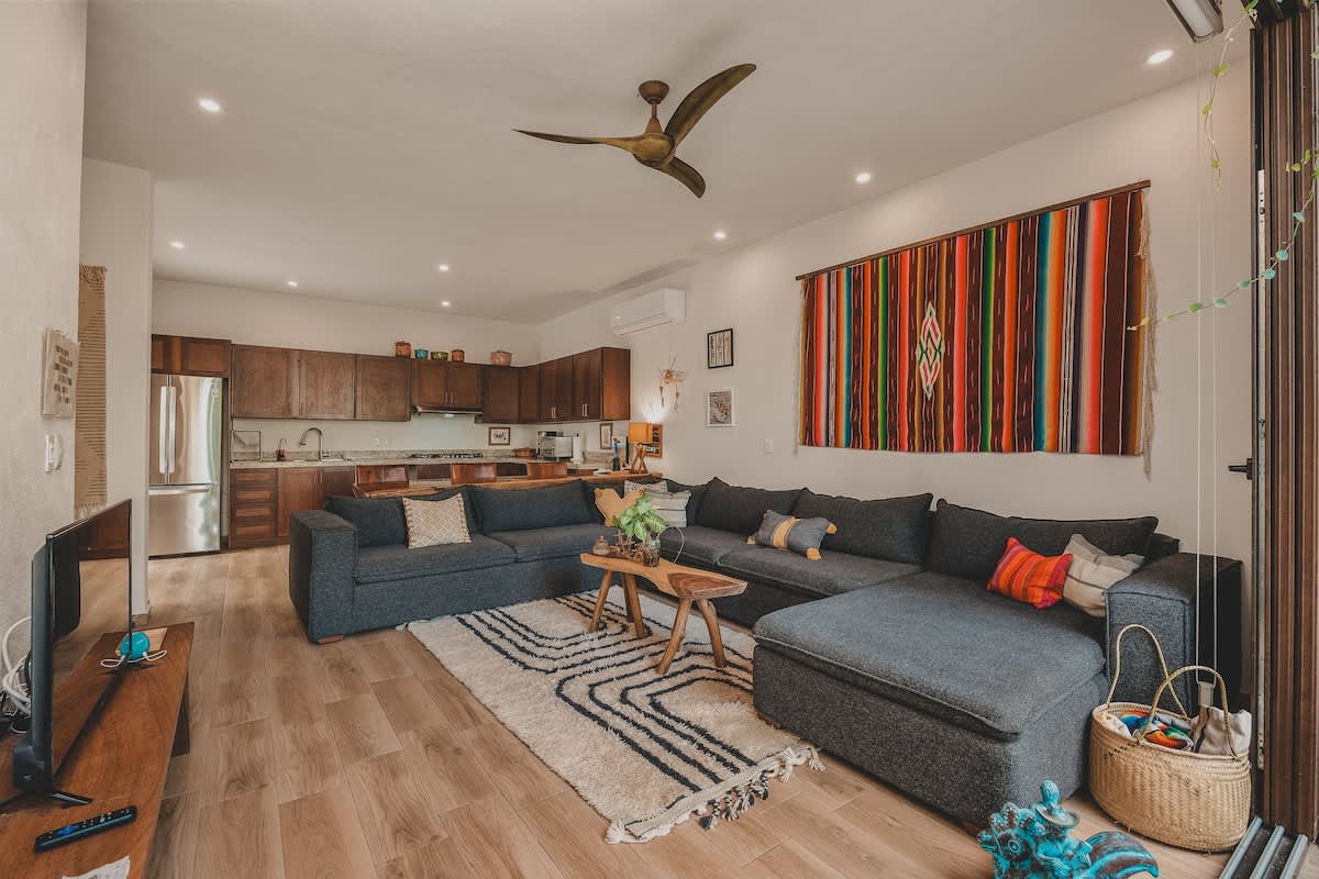 The bright living room boasts a large dark gray sectional sofa, a geometric patterned rug, and a colorful woven tapestry, with a ceiling fan and an air conditioning unit, and expansive sliding doors offering views of the lush outdoor greenery.