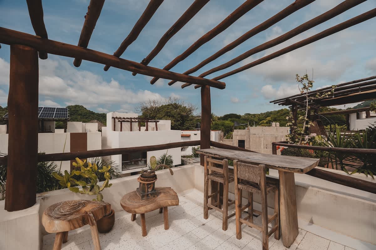 An expansive rooftop deck with a rustic wooden pergola offers a high-top bar table with two stools and a small wooden side table, overlooking a residential area with lush greenery and a cloudy sky.