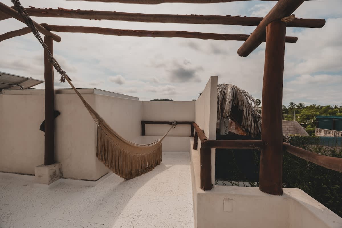 A cozy rooftop balcony features a woven hammock suspended from a rustic wooden pergola, offering a relaxing elevated outdoor space with white walls and views of distant greenery and sky.