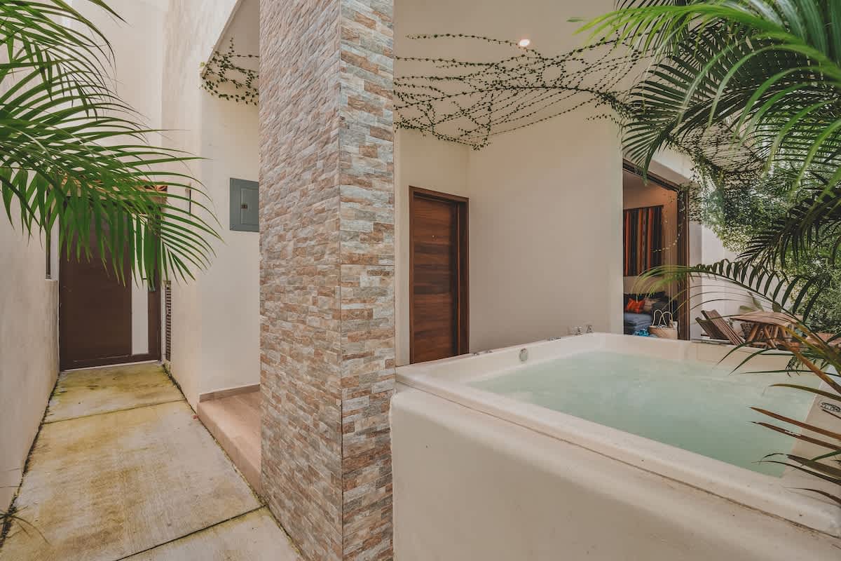 The exterior view showcases a private patio with a hot tub, a stone-tiled pillar, and a concrete pathway, all framed by lush green tropical foliage and palm trees.