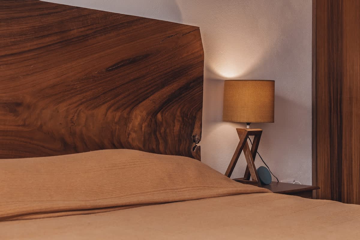 A cozy bedroom features a striking live-edge wooden headboard, a bed with soft light-colored bedding, and a bedside table with a modern lamp and a small smart speaker.
