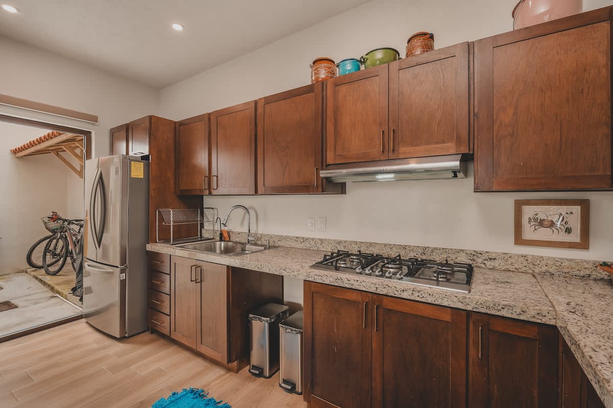 The kitchen features dark wood cabinets, granite countertops, and a stainless steel refrigerator. A double sink is visible, and a glass door opens to an outdoor patio with a bicycle, suggesting an indoor-outdoor flow.