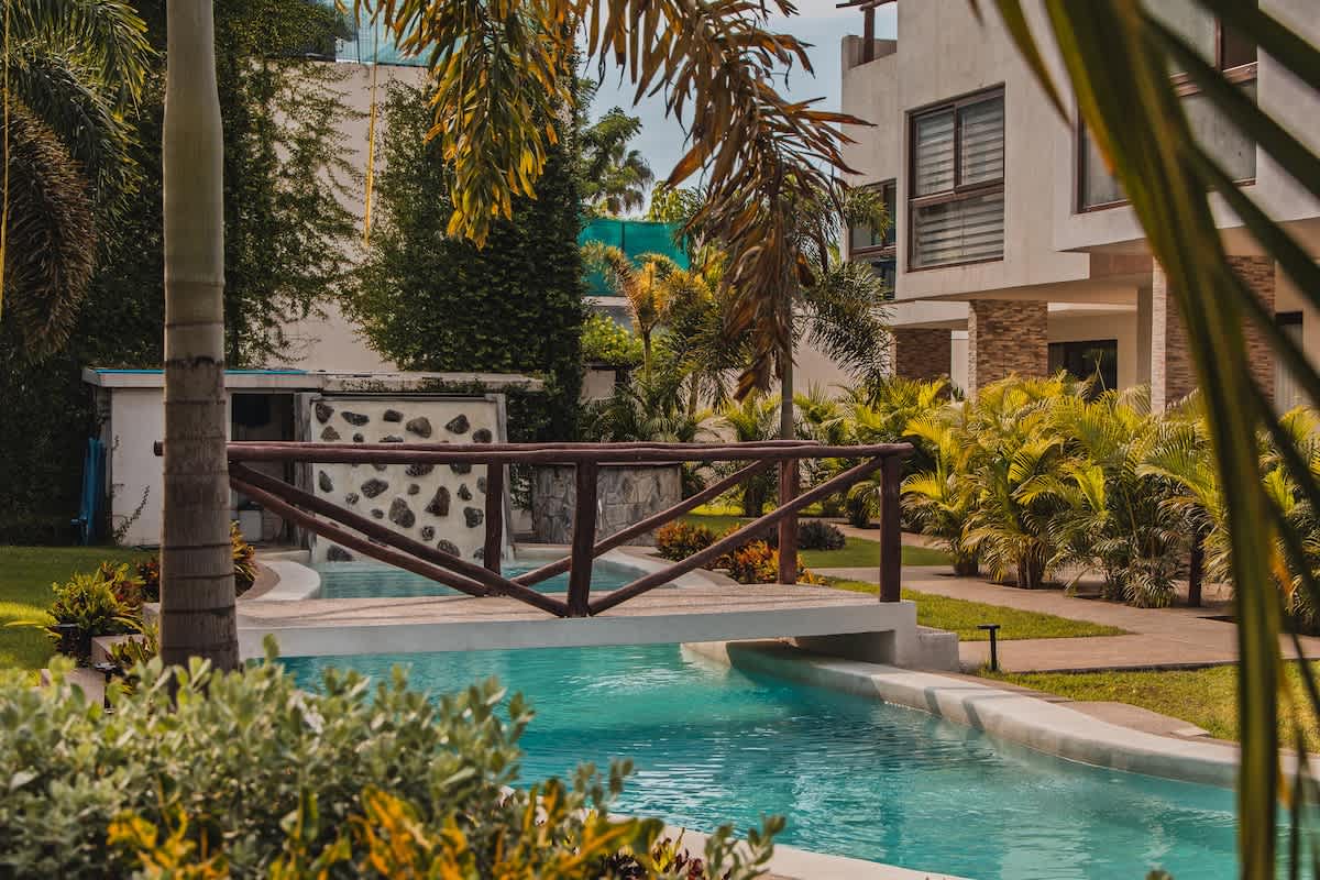 A serene outdoor scene features a winding swimming pool with a small wooden bridge crossing over it, surrounded by lush green tropical foliage and palm trees. Modern buildings are visible in the background.