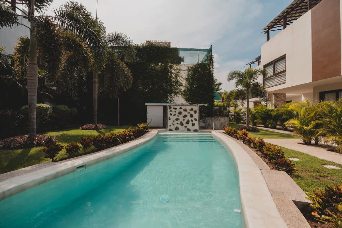 A serene, light blue swimming pool with a curved edge is surrounded by manicured green grass and vibrant tropical landscaping, leading towards modern white buildings under a partly cloudy sky.