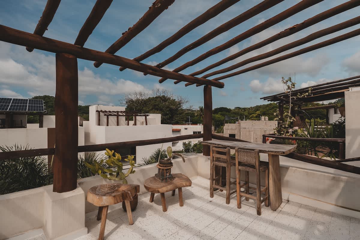 A rooftop balcony features a rustic wooden pergola providing shade over a small wooden bar table with two stools and two carved wooden side tables. The white tiled floor and surrounding lush greenery offer a relaxing outdoor space with views of other buildings.