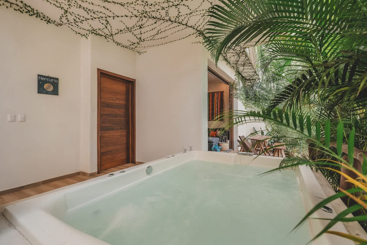 The outdoor patio features a white built-in hot tub, surrounded by vibrant green tropical plants, with an open sliding glass door revealing an inviting indoor living space.