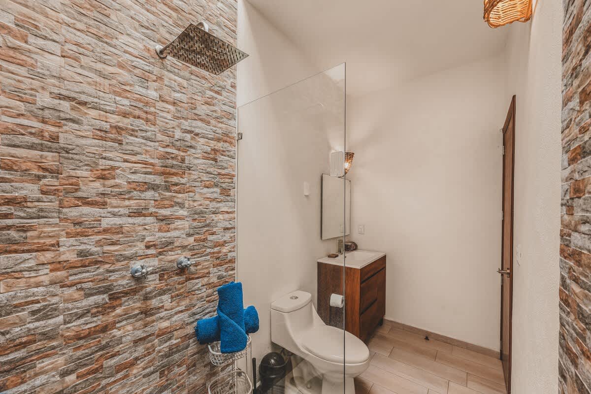Modern bathroom featuring a spacious walk-in shower with textured stone tile walls and a rain showerhead, a white toilet, and a wooden vanity with a sink and mirror. Blue towels are neatly stacked.