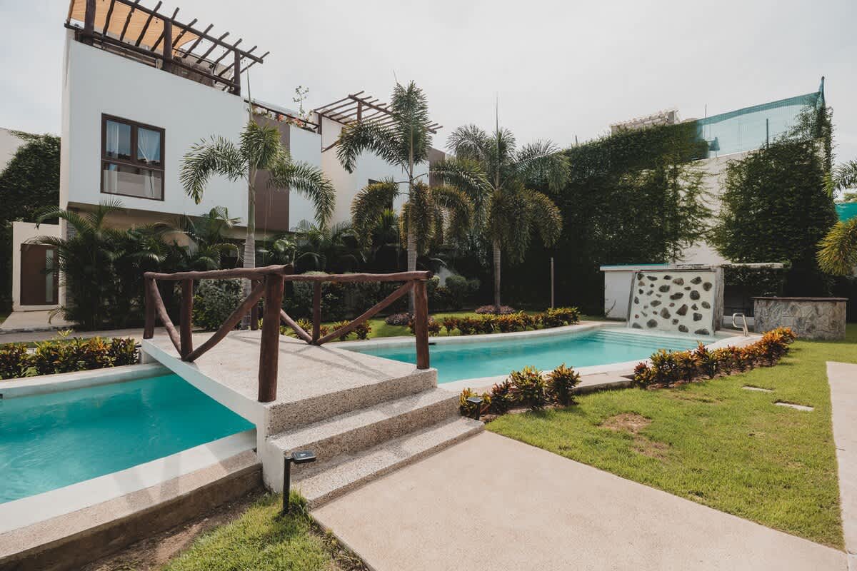A light blue swimming pool with a rock-faced waterfall feature is nestled in a vibrant green lawn, surrounded by lush tropical foliage and palm trees. A concrete pathway leads to a small wooden bridge spanning a narrow section of the pool.
