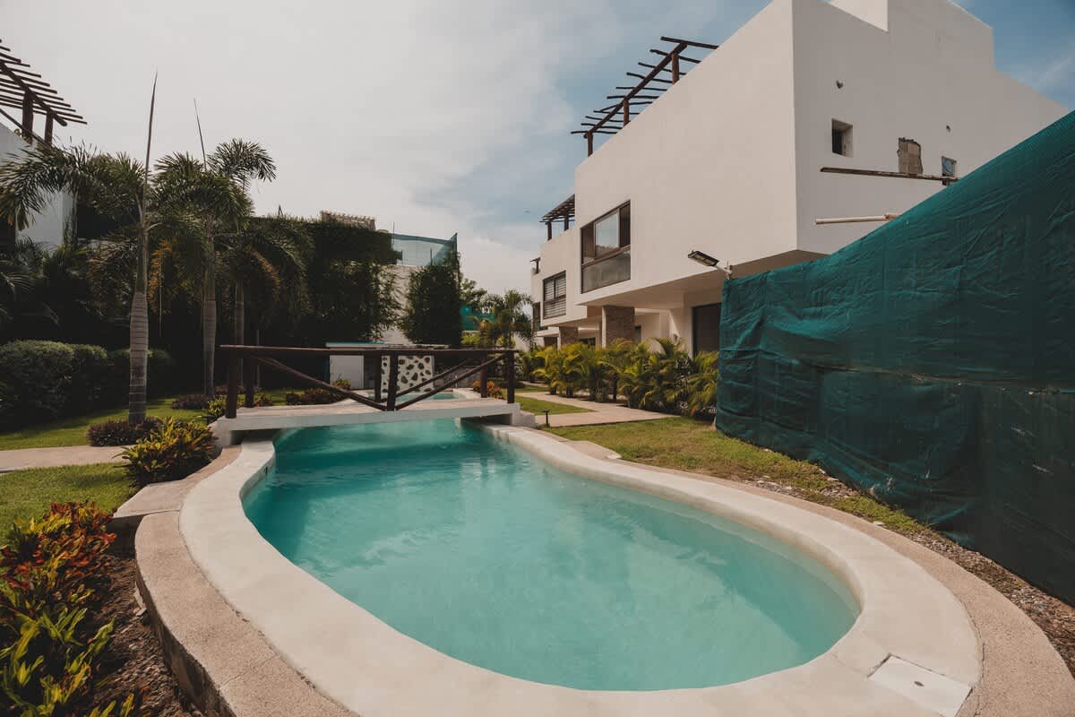 A serene oval-shaped light blue swimming pool is surrounded by a well-kept green lawn and vibrant tropical plants. Modern white buildings with dark window frames are visible in the background, along with a wooden bridge over a connecting water channel.