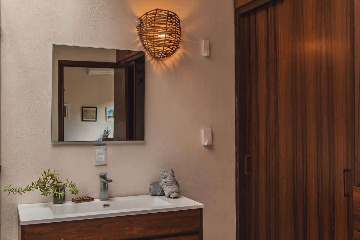 A well-lit bathroom features a white vanity with a chrome faucet, a large mirror reflecting an adjacent room, a rustic woven wall sconce, and a dark wood sliding closet door.