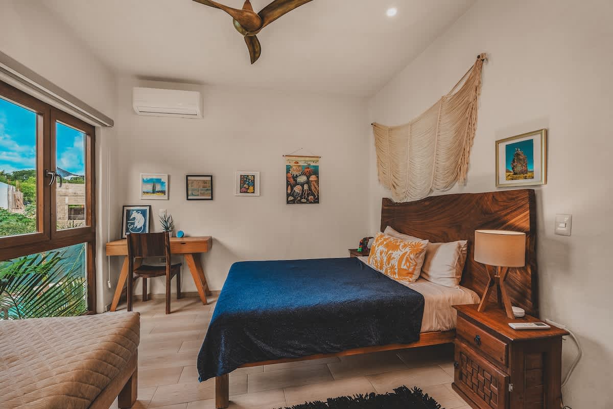Bright bedroom with a large bed featuring a distinctive wooden headboard, a wooden desk by the window, a daybed, and various artworks adorning the light-colored walls. A ceiling fan is visible overhead.