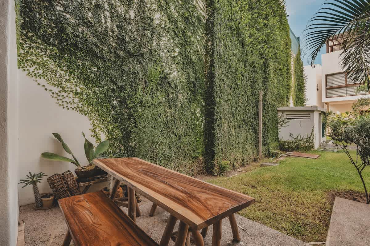 A spacious green yard offers a long, rustic wooden dining table with benches, bordered by a tall wall completely covered in dense green vines and a small utility shed.