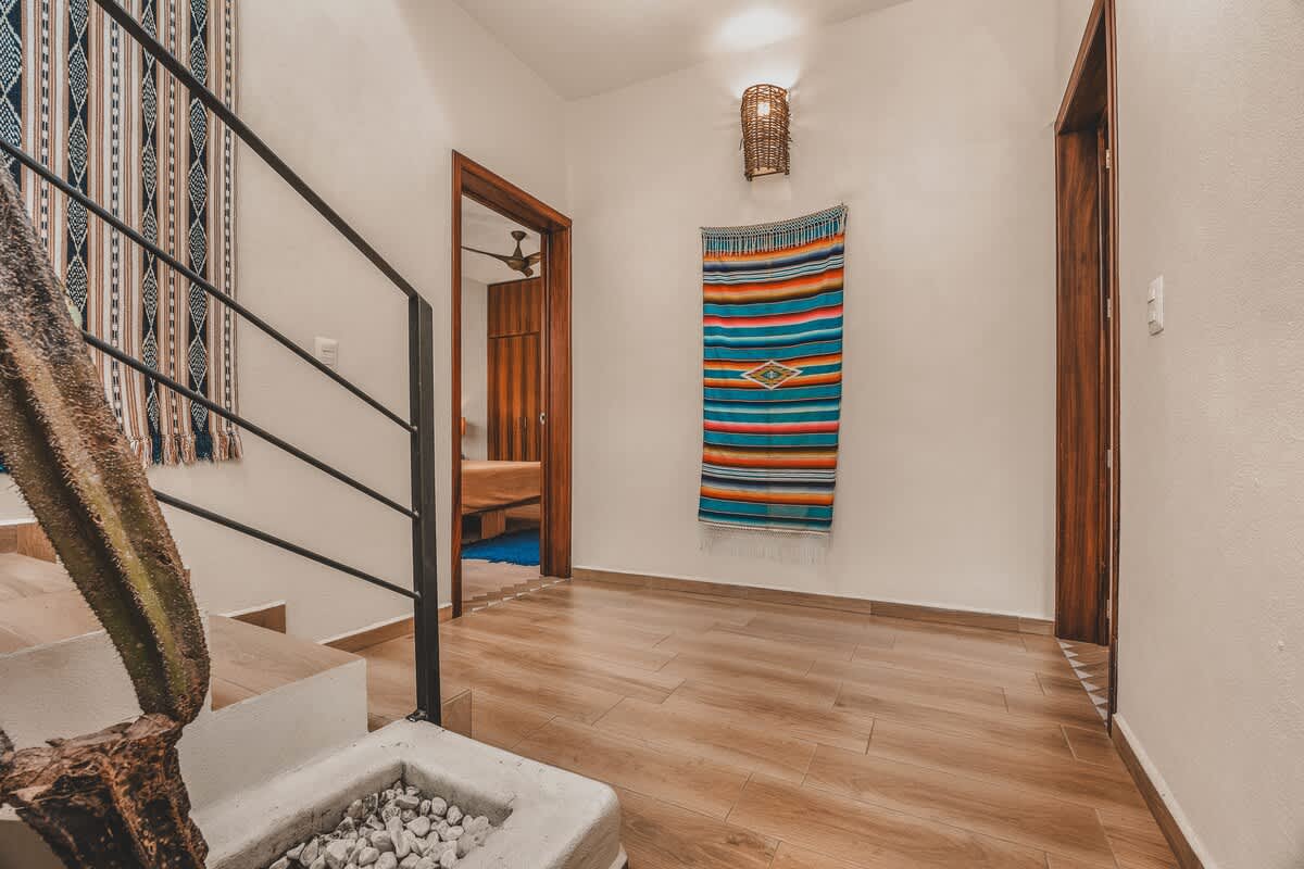 Bright hallway with light wood-look tile flooring, featuring a colorful woven tapestry on the wall, a doorway leading to a bedroom, and a view of the staircase with a cactus plant below.