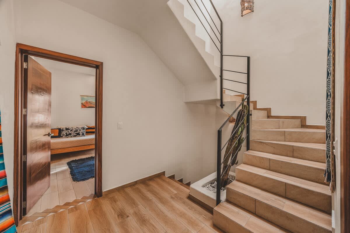 A bright interior hallway features a staircase with light-colored tiled steps and a black metal railing, leading up from a landing with wood-look flooring, with a doorway to another room visible.