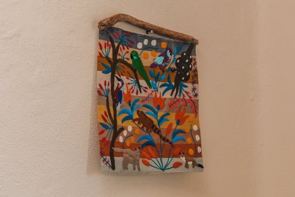 A colorful embroidered textile wall hanging, suspended from a natural wood branch, depicts various animals like an iguana, birds, and foxes amidst vibrant foliage and abstract shapes.