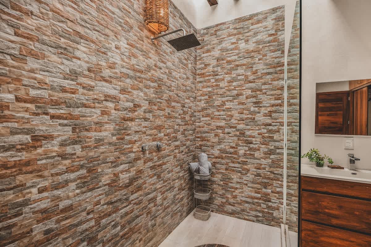 A detailed view of the spacious walk-in shower, featuring a rain shower head, a stacked stone accent wall in earthy tones, and a circular wooden inlay on the light tile floor.