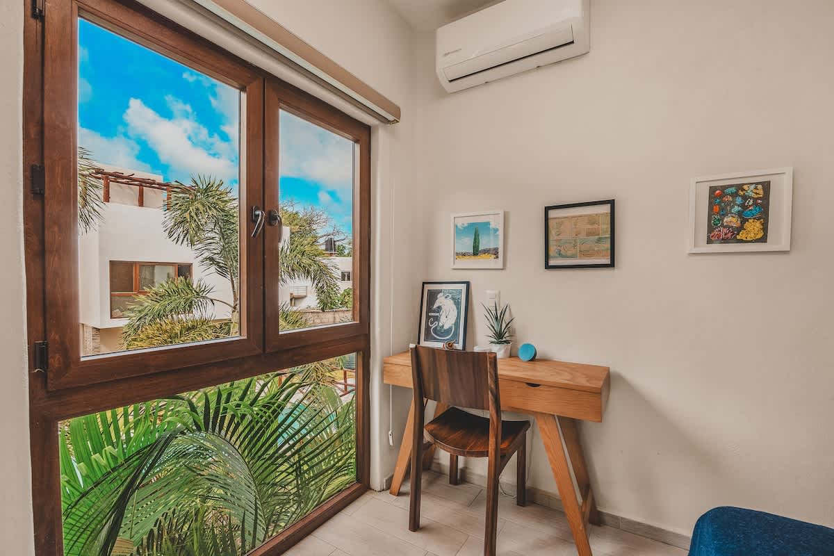 Detailed view of a wooden desk and chair next to a large window, offering a vibrant view of blue sky and palm trees, with framed artwork adorning the wall above the desk. A small plant sits on the desk.
