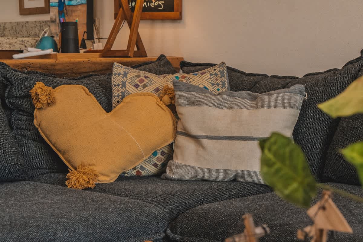 A close-up of a dark grey textured sofa adorned with decorative pillows, including a yellow heart-shaped pillow with tassels and a striped rectangular pillow with geometric patterns.