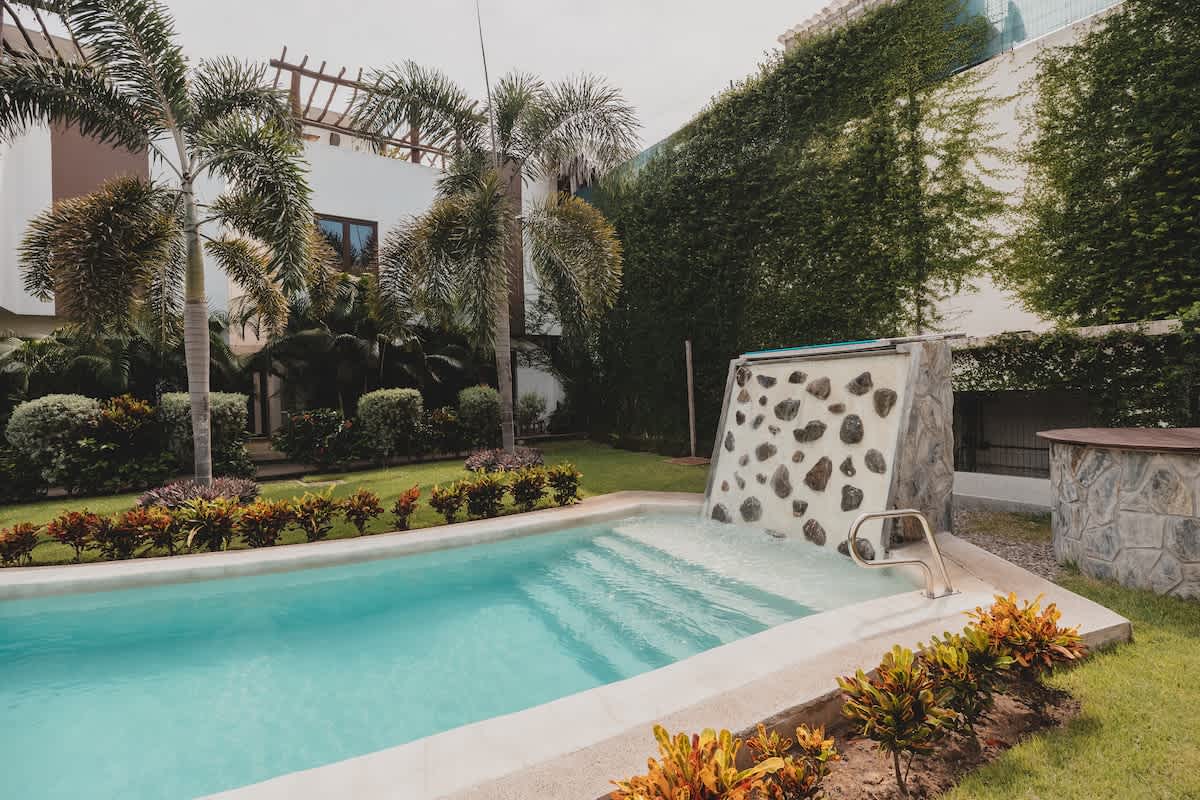 A kidney-shaped swimming pool with light blue water and a rock-faced waterfall feature is set in a green lawn, surrounded by tropical plants. Modern white buildings with lush green ivy on one wall are visible in the background.