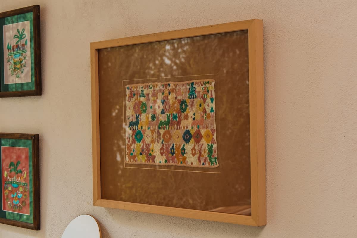 A framed piece of colorful textile art with intricate patterns and animal motifs is displayed on a light-colored wall, adding texture and cultural flair to the room.