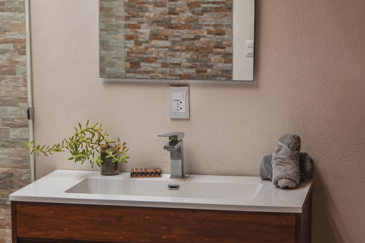 A bright bathroom vanity features a white rectangular sink, a modern chrome faucet, a small potted plant, and neatly rolled grey towels, all beneath a large wall mirror.