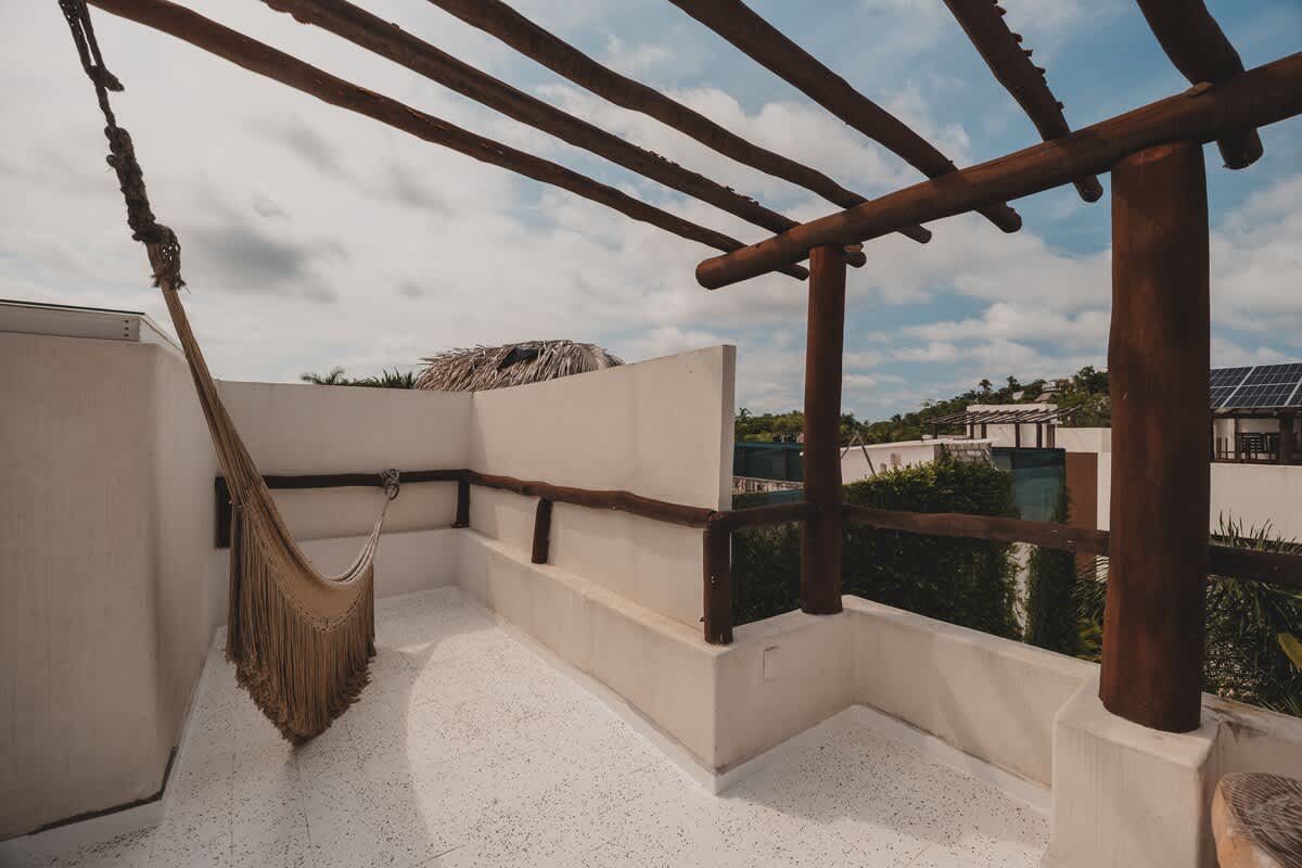 An elevated rooftop balcony with a white floor and walls is shaded by a rustic wooden pergola, featuring a woven hammock and offering expansive views of the surrounding tropical landscape and sky.