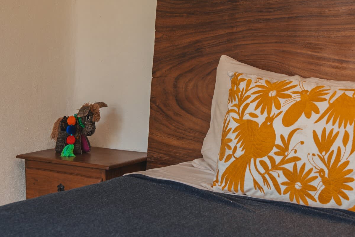 A bed with a dark blue blanket and a live-edge wood headboard is accented by a white pillow featuring a vibrant yellow bird and floral embroidery, next to a wooden nightstand with a decorative donkey figurine.