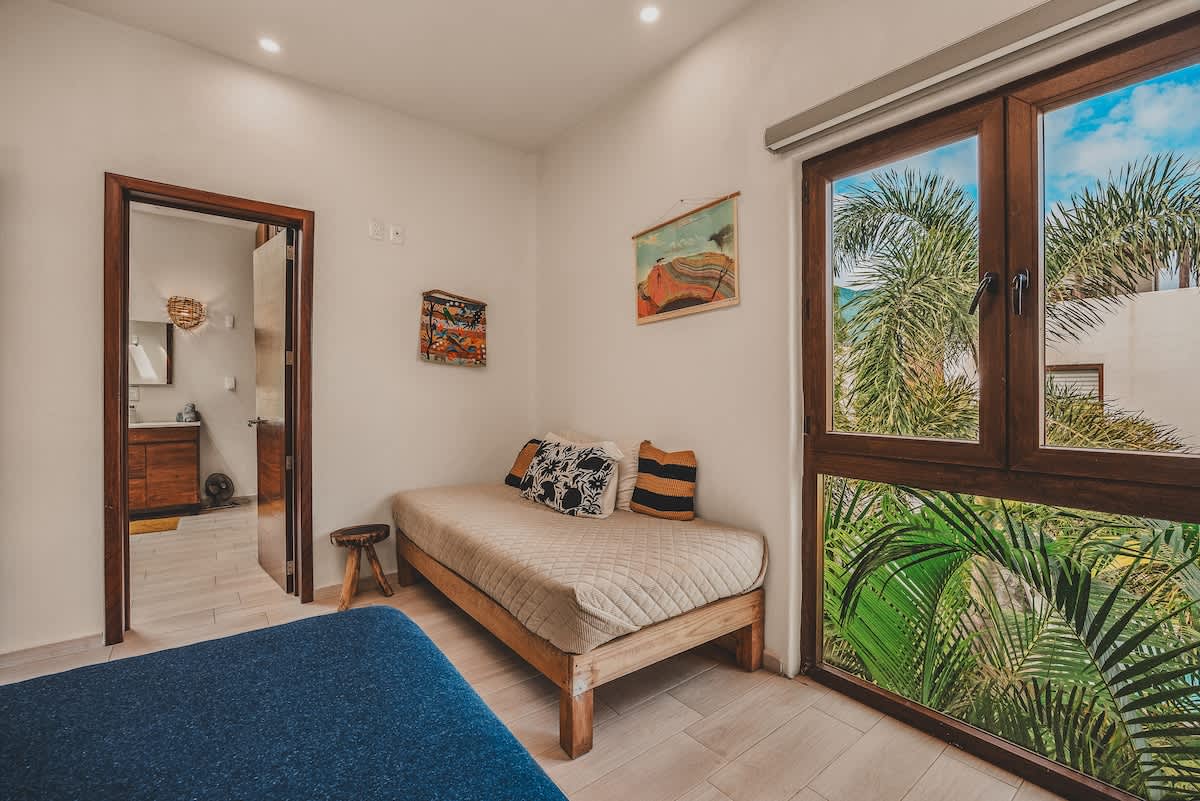 A bright bedroom featuring a daybed with patterned pillows, light tile flooring, and a large window offering lush tropical views of palm trees. A dark wood doorway leads to an adjacent bathroom.