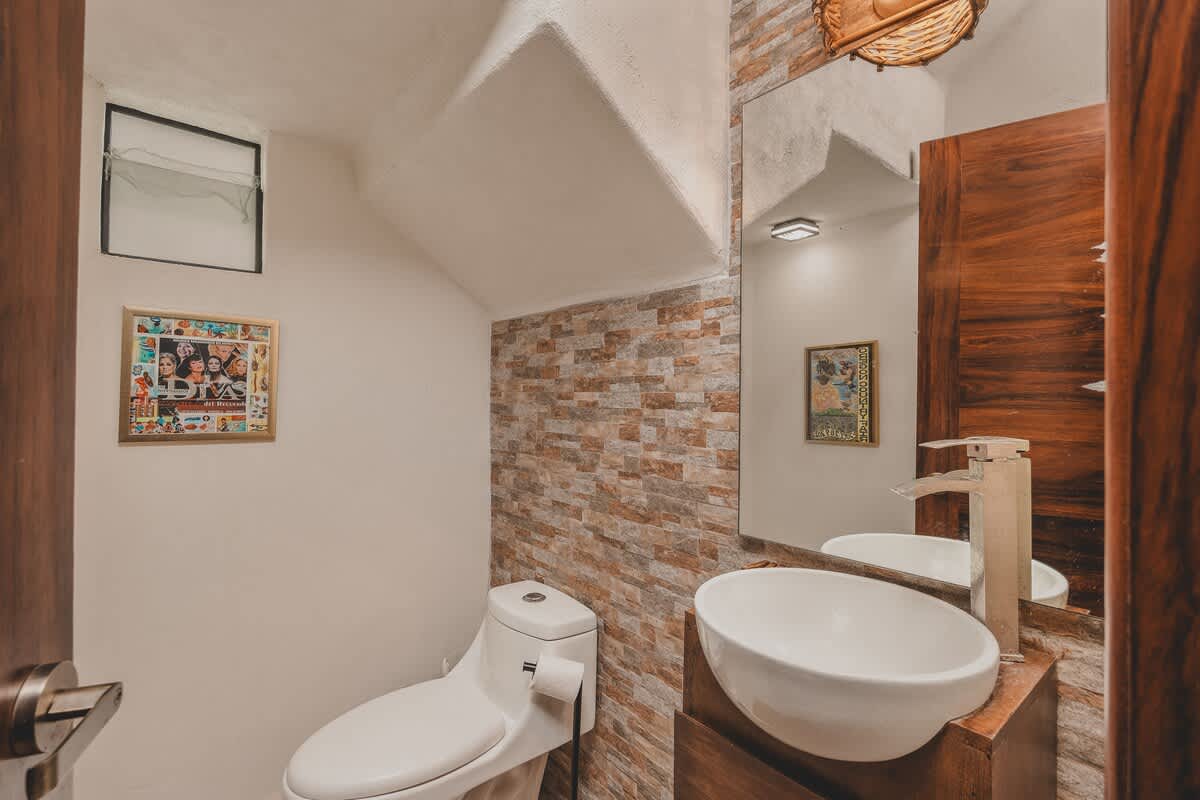 A compact bathroom with a white toilet, a modern vessel sink on a dark wood vanity, and a mirror, with a stacked stone accent wall behind the sink and a woven light fixture.