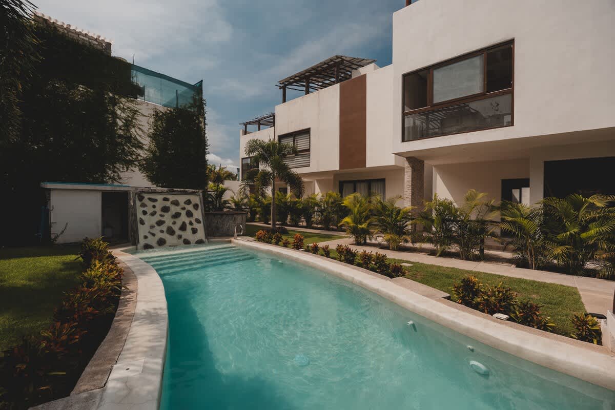 A clear, light blue swimming pool with integrated steps is bordered by a concrete edge and lush green landscaping, with a decorative stone-patterned wall feature at one end and modern buildings beyond.