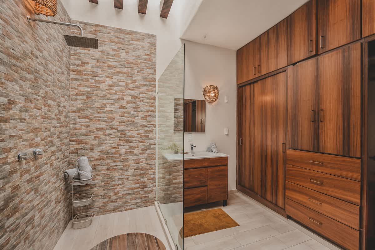 A wide shot of a contemporary bathroom showcasing a glass-enclosed walk-in shower with a stacked stone wall, a dark wood vanity, and an extensive built-in wooden wardrobe, under a ceiling with exposed beams.