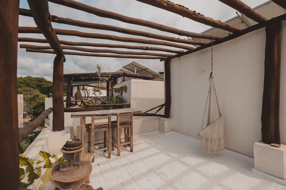 A rustic rooftop deck features a wooden pergola, a high-top bar table with two stools, and a macrame hanging chair, all set against a white wall and a cloudy sky. Solar panels are visible in the background.