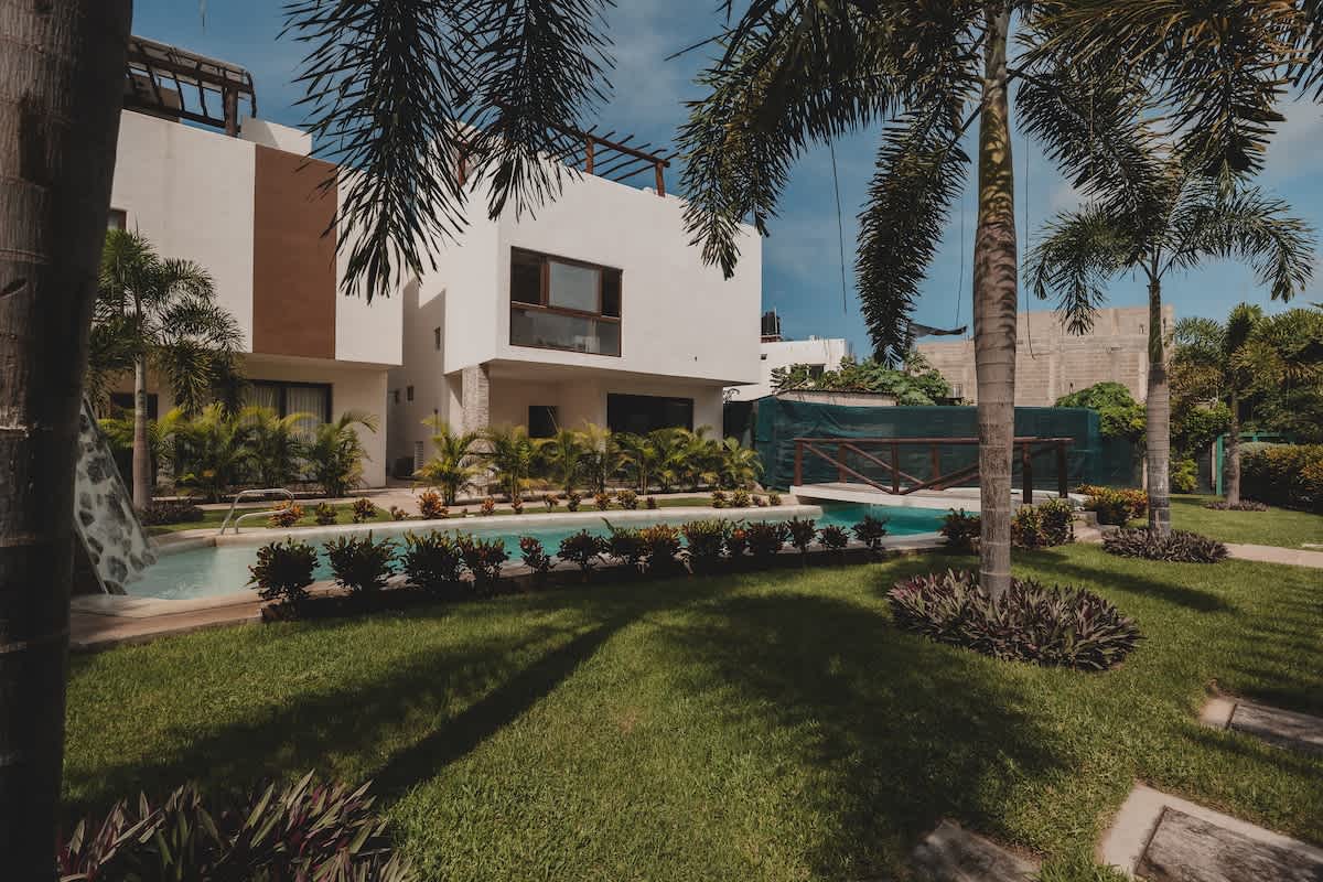 A spacious green lawn and a curved swimming pool are central to this outdoor area, flanked by contemporary white buildings with brown accents and surrounded by various tropical plants and palm trees.