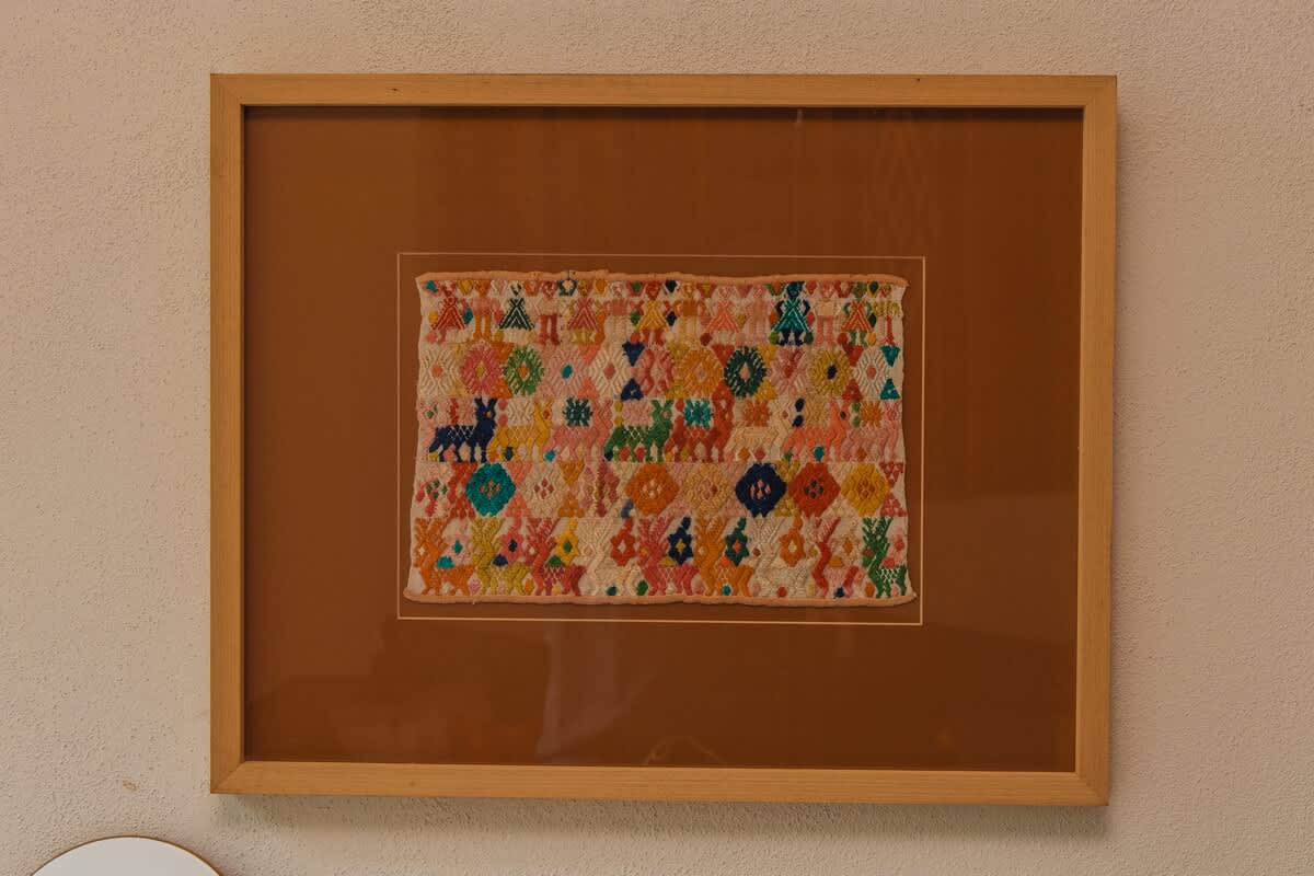 A framed piece of colorful textile art displays a vibrant array of embroidered geometric shapes and animal figures in shades of pink, blue, green, and yellow, set against a light background.