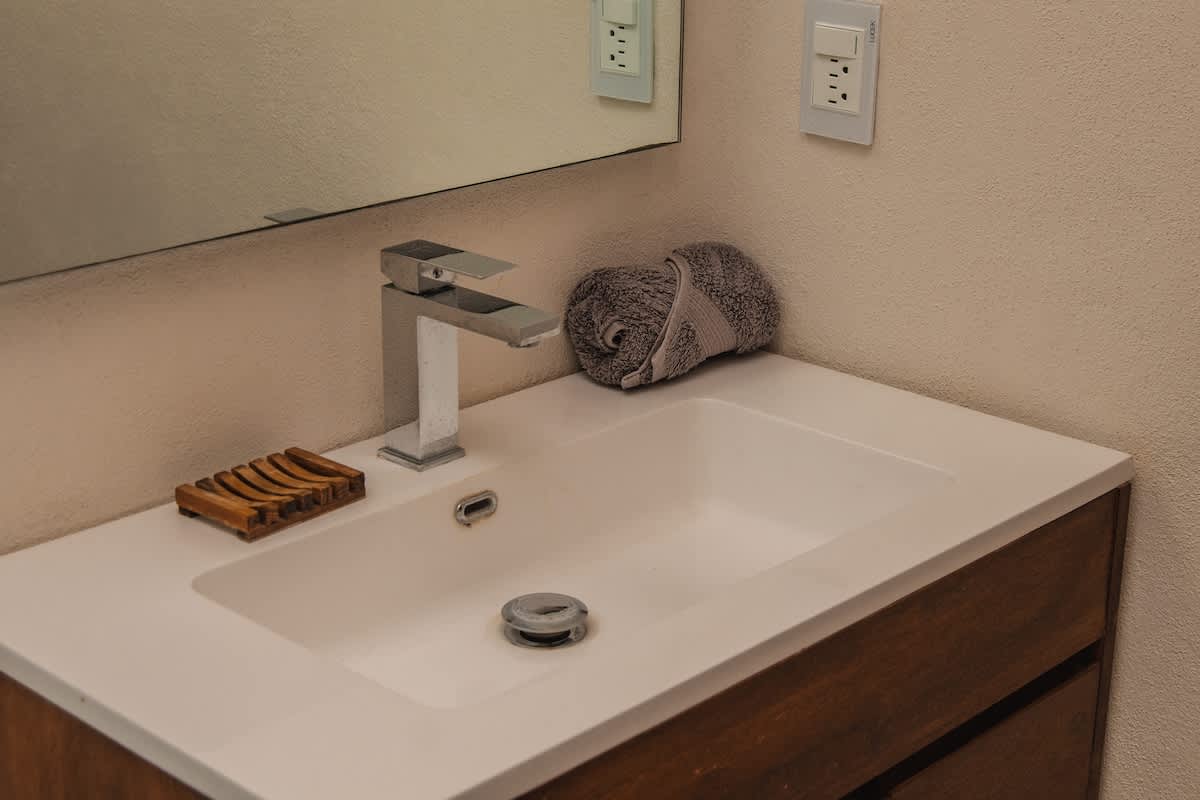 A clean bathroom vanity features a white rectangular sink, a modern chrome faucet, a wooden soap dish, and a neatly rolled grey towel, all beneath a large wall mirror.