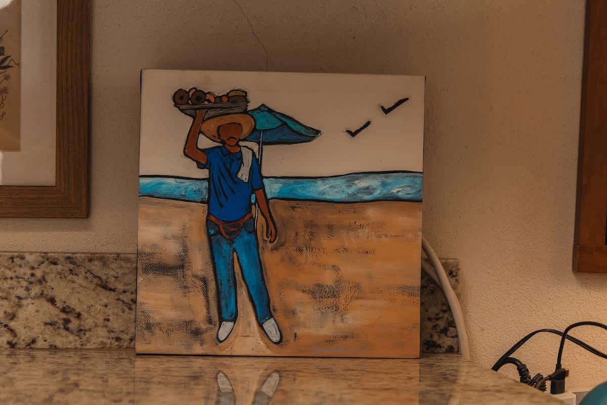 A vibrant painting on a square canvas depicts a person in a hat carrying a tray of goods on a sandy beach with blue water and sky, resting on a speckled granite countertop.