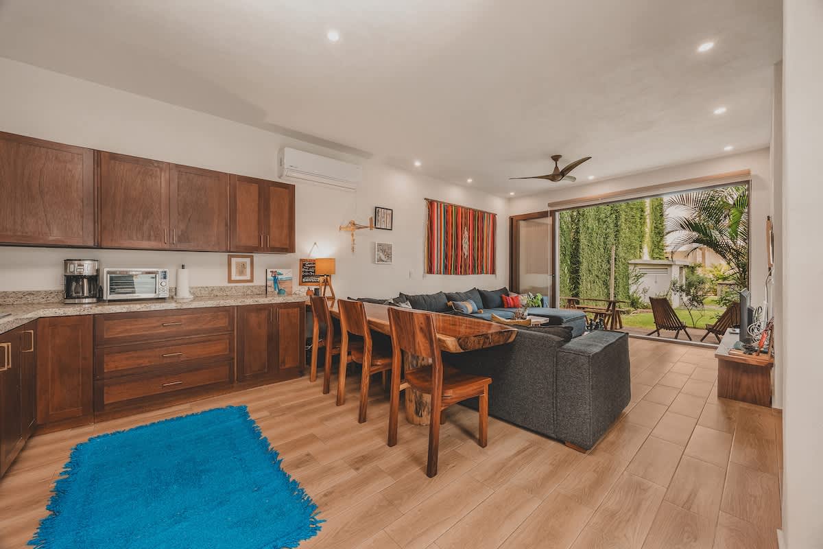 An open-concept living space features a dark gray sectional sofa, a patterned rug, and a vibrant tapestry on the wall, seamlessly connecting to a kitchen with dark wood cabinetry and granite countertops, with large sliding doors opening to the outdoors.
