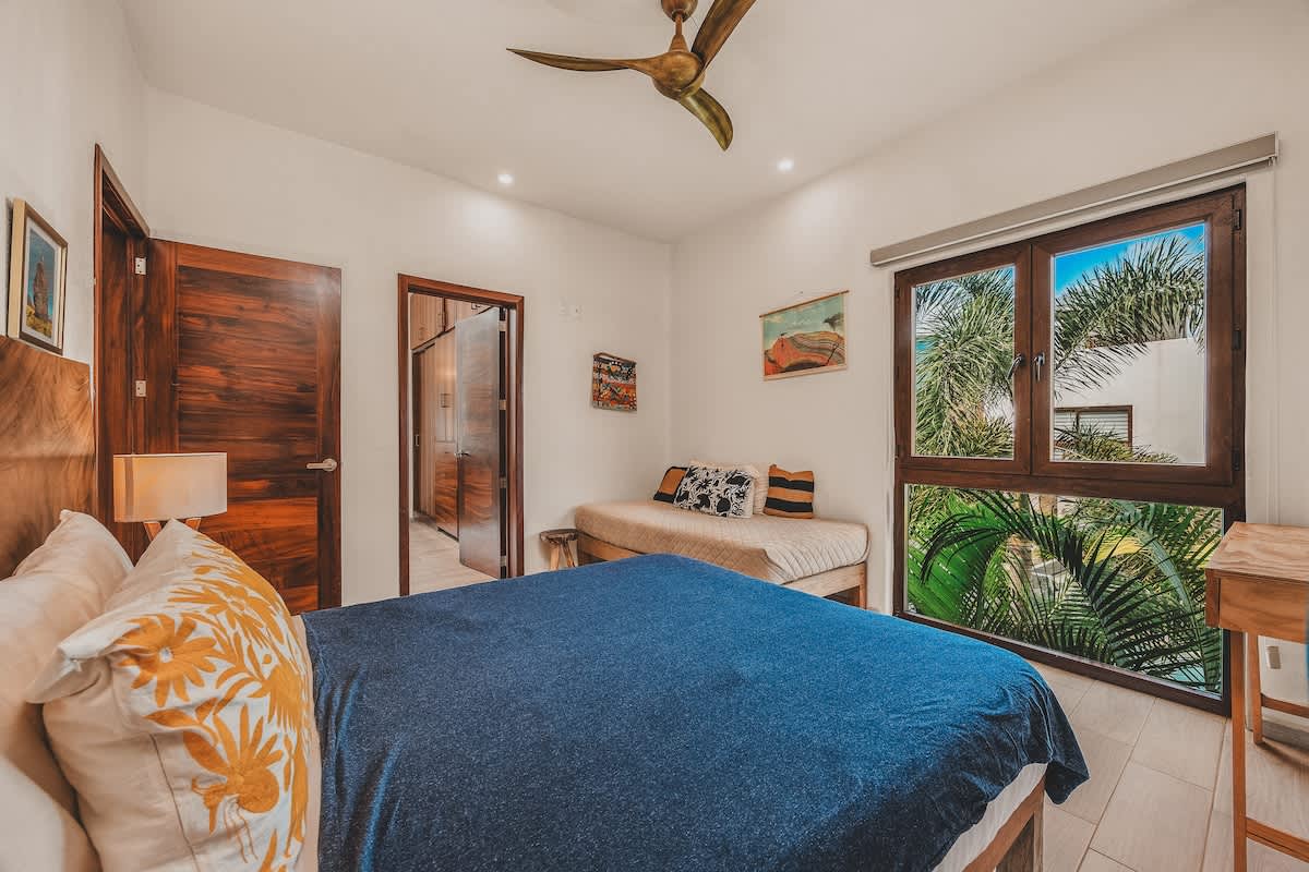 Inviting bedroom featuring a large bed with a natural wood headboard, a daybed, and a wide window offering views of lush tropical foliage, with a ceiling fan overhead. Artwork and wooden doors are visible.