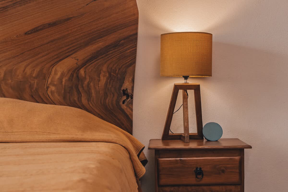 A cozy bedroom corner features a bed with a warm-toned blanket, a live-edge wood headboard, and a wooden nightstand with a modern lamp and a small blue smart speaker.