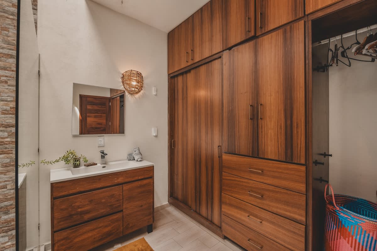 A bathroom featuring a dark wood vanity with a white sink and mirror, complemented by a large built-in wooden wardrobe with sliding doors and drawers, all set against light-colored walls and tile flooring.