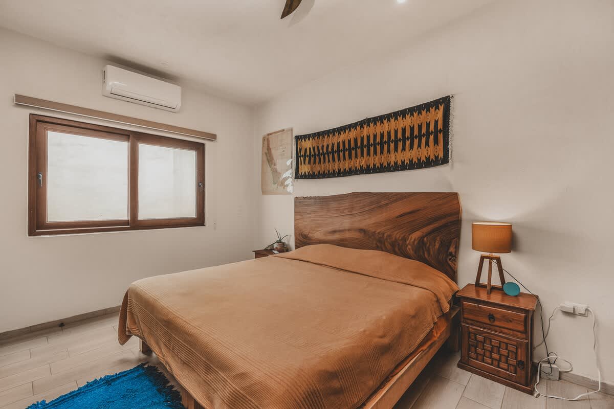 A bright bedroom features a large bed with a live-edge wooden headboard, a wooden nightstand with a lamp, and a blue rug on light wood-look tile floors, with a window and an air conditioning unit above it.