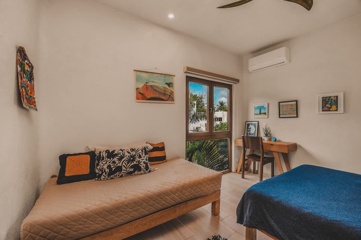 Well-lit bedroom showcasing a wooden desk and chair positioned by a large window with views of tropical foliage, alongside a daybed and various framed artworks on the wall. An AC unit is mounted above the window.