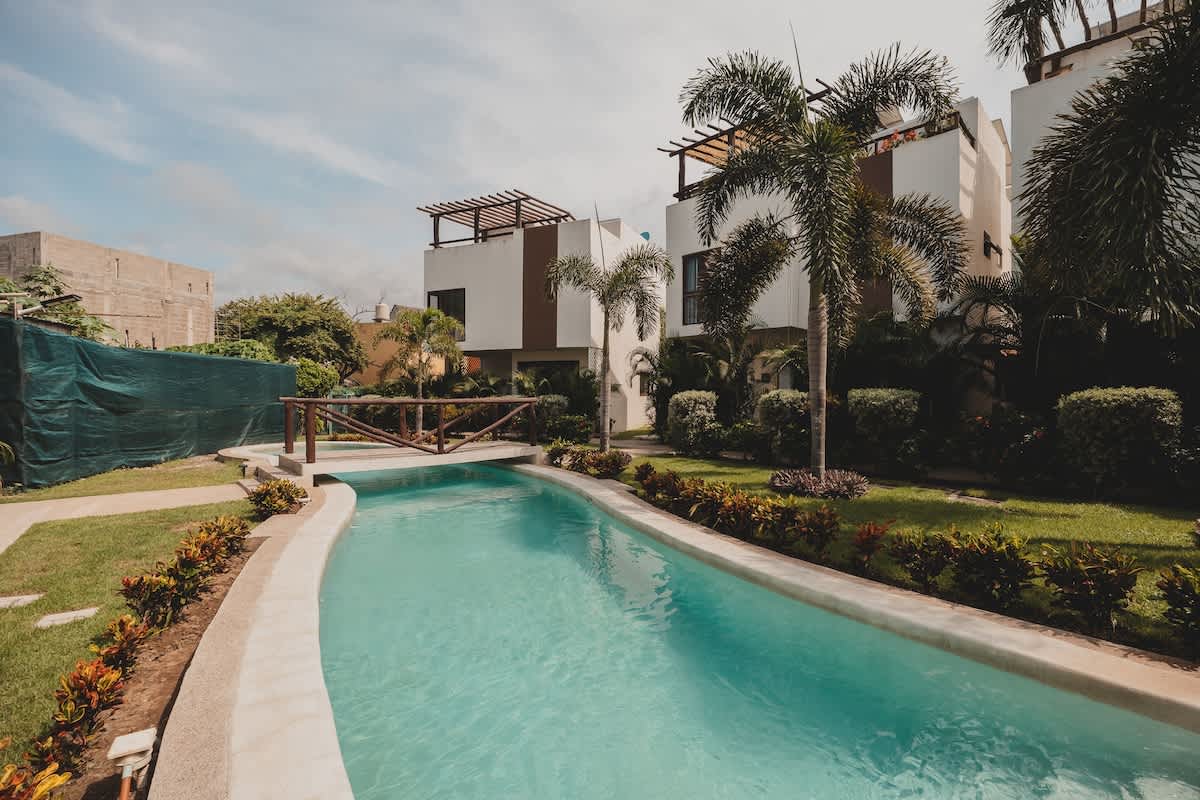 The curved, light blue swimming pool extends through a landscaped garden, featuring a small wooden bridge crossing over it, with modern white buildings and lush greenery in the background.
