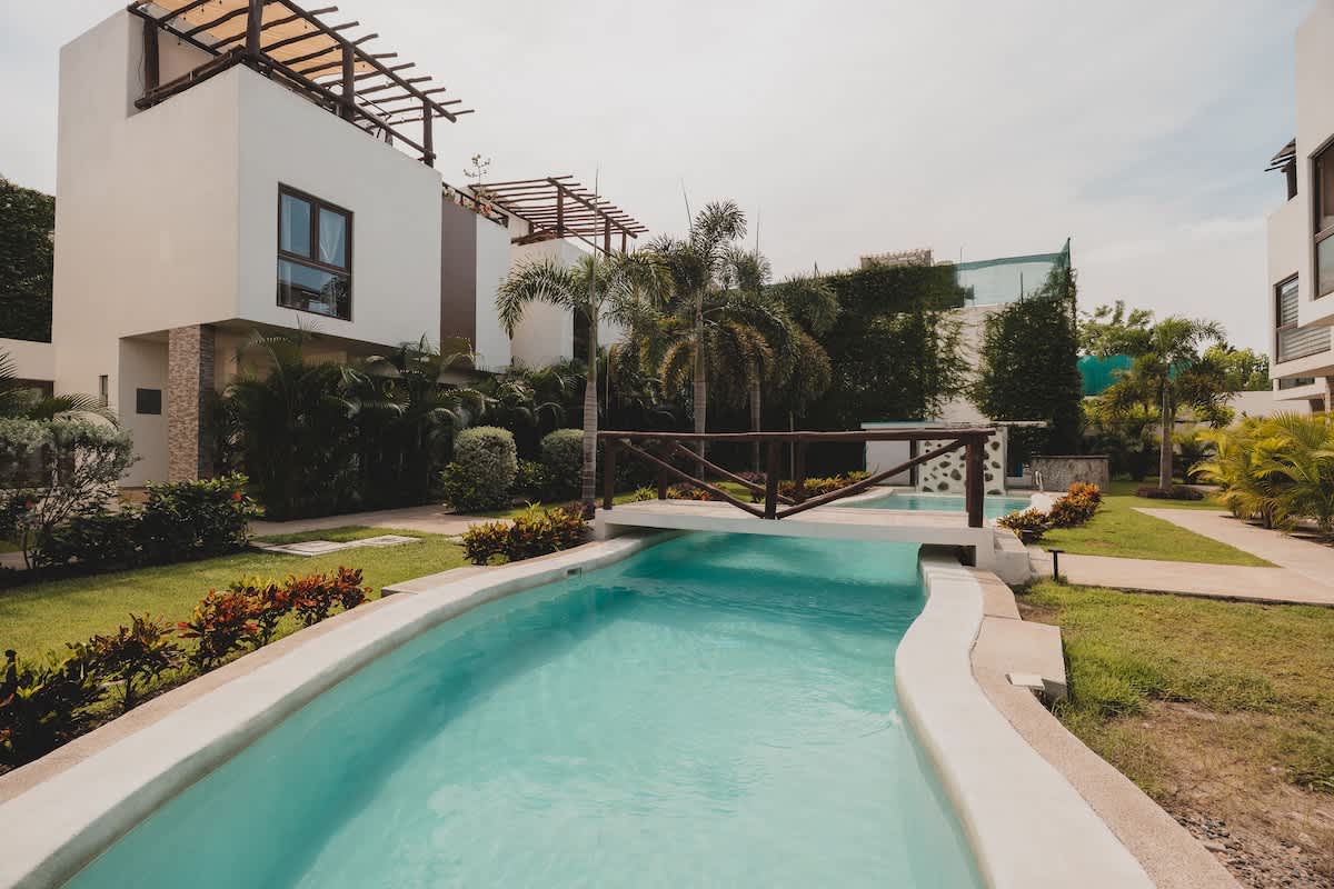 A long, narrow light blue swimming pool winds through a well-maintained green lawn, bordered by tropical plants and palm trees. A small wooden bridge with concrete steps crosses over the pool, connecting different sections of the property.