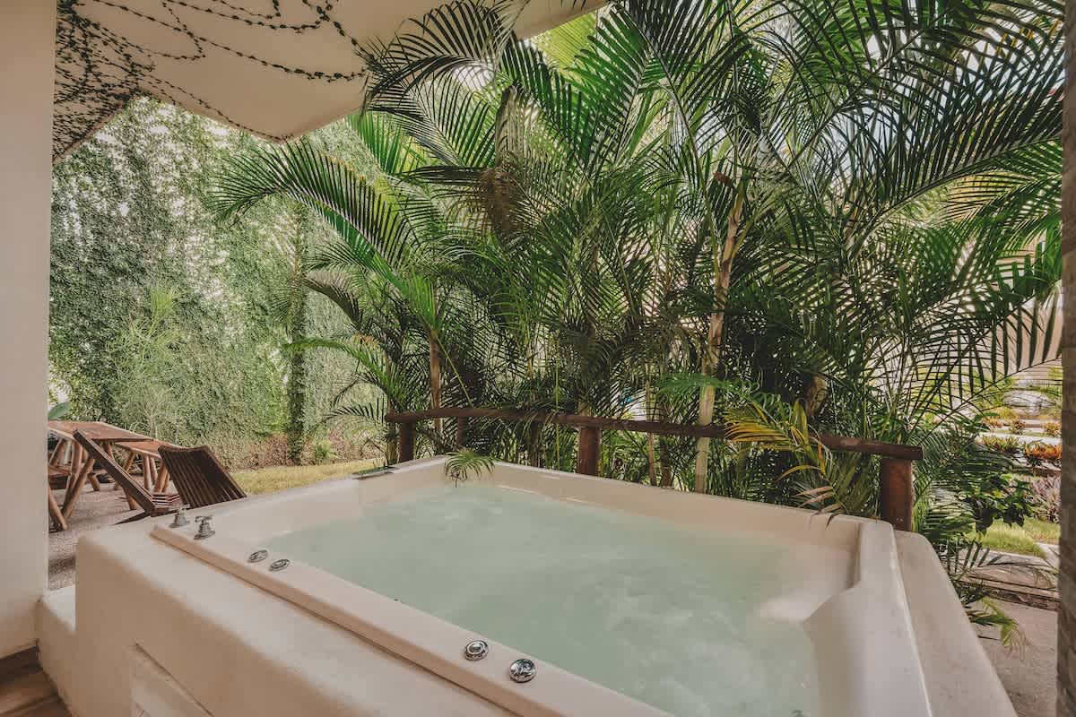 A white built-in hot tub with bubbling water is set on a patio, surrounded by vibrant green tropical plants and palm trees, creating a private, serene outdoor oasis. Wooden lounge chairs are visible in the background.