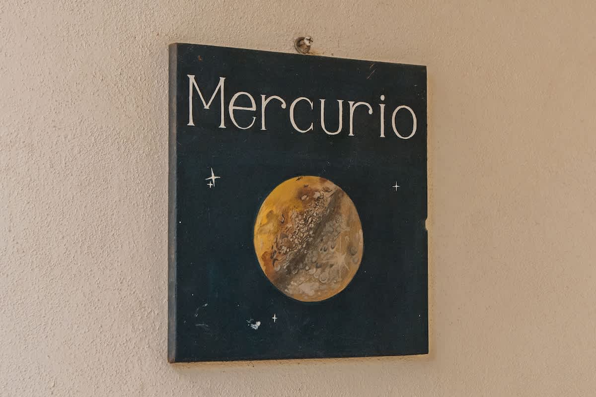 A square, dark blue decorative sign with 'Mercurio' in white text and a detailed illustration of the planet Mercury, featuring craters and stars, mounted on a textured, light beige stucco wall.