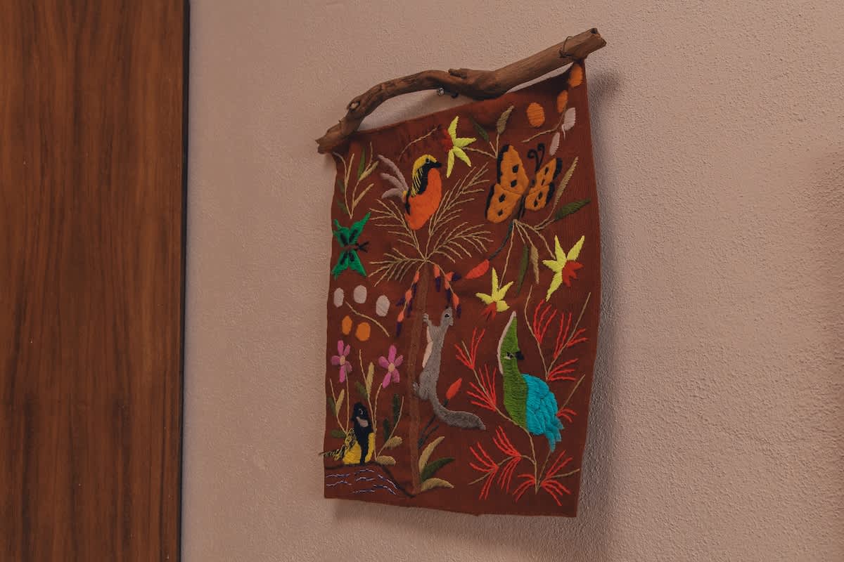 A vibrant, embroidered textile wall hanging features a natural wood branch hanger and depicts various colorful animals and plants on a brown background, adding a touch of folk art to the space.