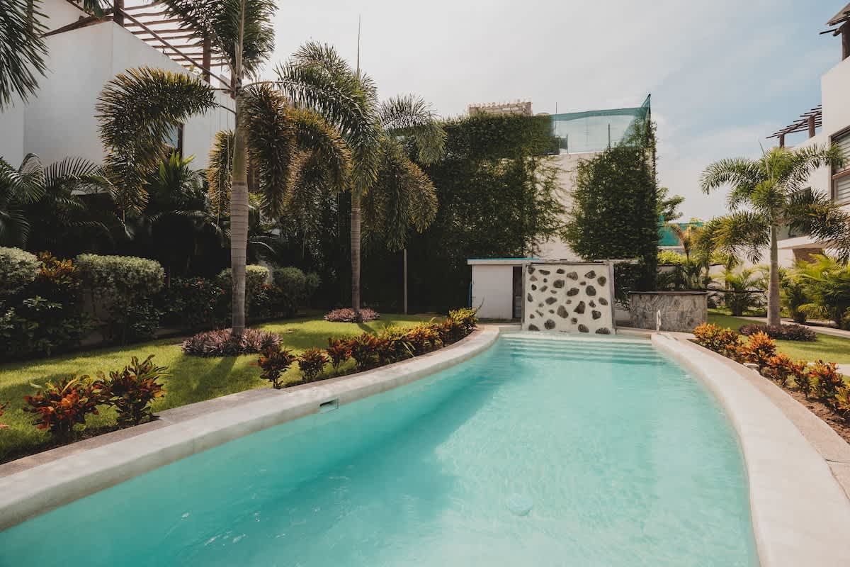 A long, winding swimming pool with clear blue water is set amidst a vibrant green lawn and dense tropical foliage, including tall palm trees, with a modern building partially visible in the distance.