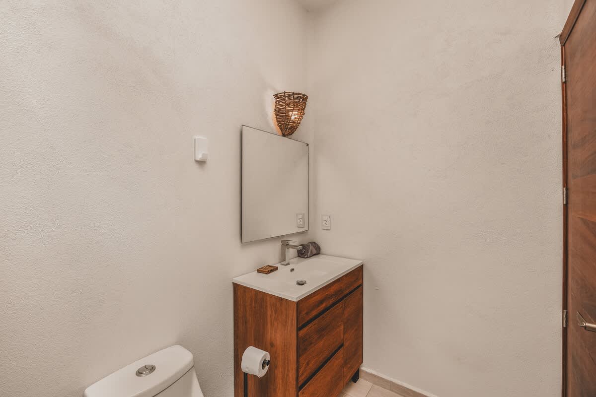 A clean bathroom features a wooden vanity with a rectangular sink and a large mirror, a white toilet, and a woven wall sconce, with a dark wooden door visible on the right.