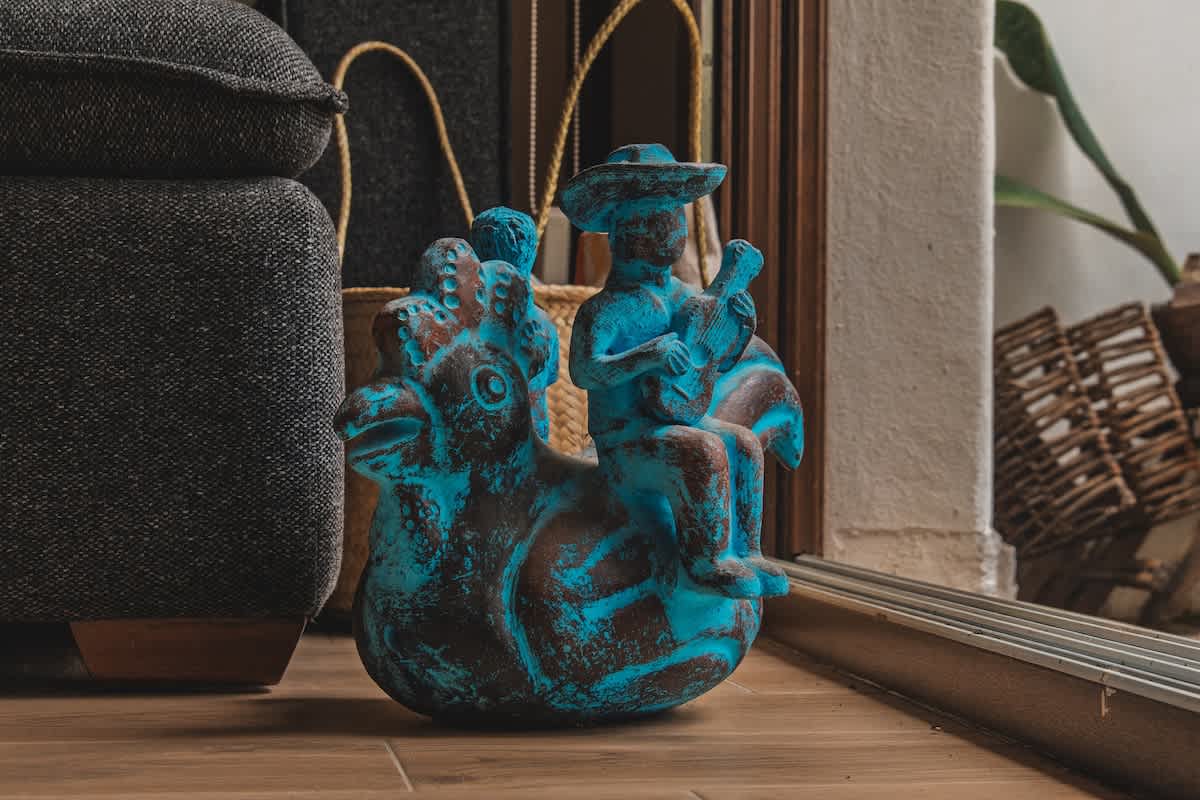 A vibrant turquoise and brown statue depicts a figure playing a stringed instrument while riding a stylized animal, placed on a light wood floor next to a dark grey upholstered furniture piece.